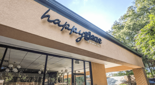 Sign for "happyplace DONUTS" on a building exterior with trees in the background and a clear blue sky.