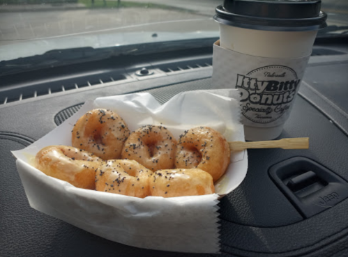 A plate of glazed donuts with poppy seeds next to a coffee cup from Itty Bitty Donuts on a car dashboard.