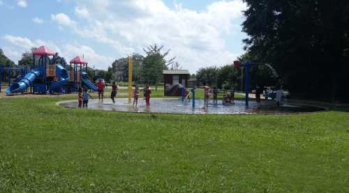 Children playing in a splash pad at a park, with playground equipment and blue skies in the background.