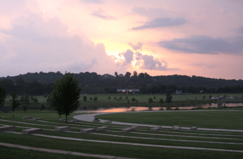 A serene landscape at sunset, featuring a grassy area, a calm river, and distant hills under a colorful sky.