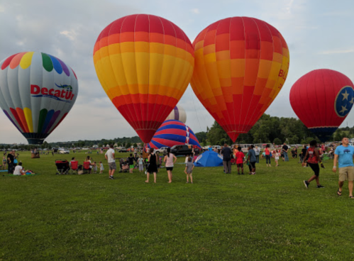 Colorful hot air balloons in various shapes and patterns at a festival, with people gathered on a grassy field.