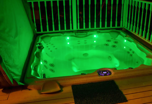 A lit hot tub with green lights, surrounded by a wooden deck and railing, set in a nighttime environment.
