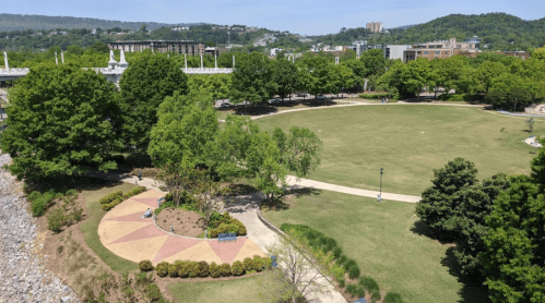A spacious park with green grass, trees, and walking paths, set against a backdrop of hills and buildings.