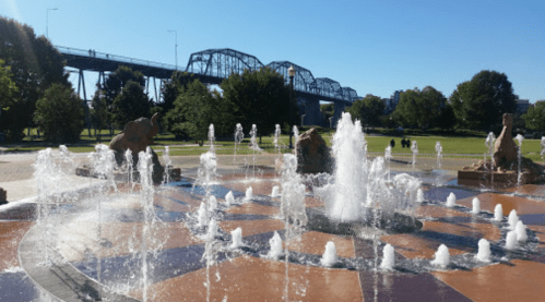 A fountain with water jets in a park, featuring sculptures and a bridge in the background under a clear blue sky.