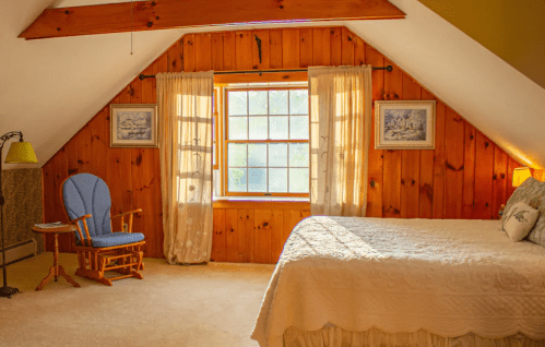 Cozy attic bedroom with wooden walls, a window, a rocking chair, and soft lighting.