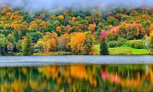 A serene lake reflecting vibrant autumn foliage in shades of orange, red, and green, surrounded by misty mountains.