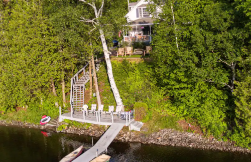 A wooden dock leads to a house surrounded by trees, with seating and kayaks by the water's edge.