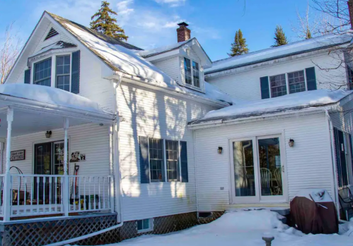 A white house with blue shutters, surrounded by snow, featuring a porch and large windows under a clear blue sky.