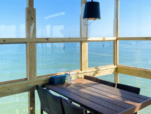 A wooden table with chairs by a large window overlooking calm blue waters and a clear sky.