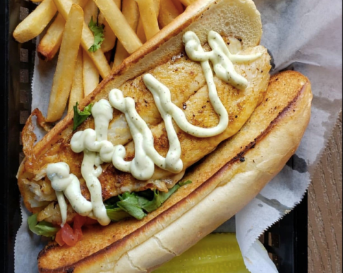 A grilled fish sandwich with lettuce and tomato, drizzled with sauce, served with fries and a pickle on the side.