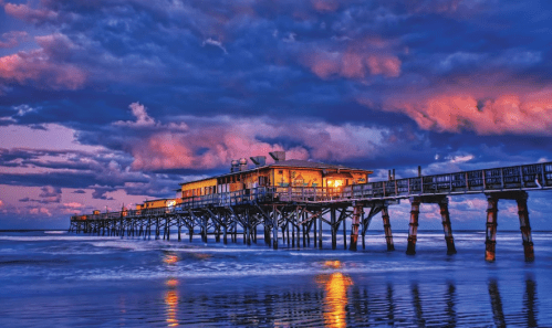 A vibrant pier at sunset, with colorful clouds reflecting on the water and warm lights glowing from the building.