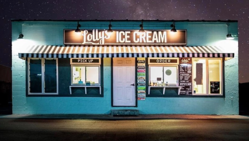 A brightly lit ice cream shop with a teal exterior, striped awning, and windows displaying menu items at night.