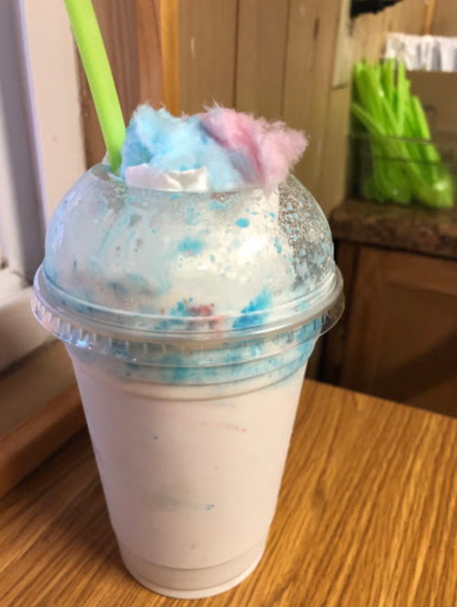 A frosty milkshake topped with blue and pink cotton candy in a clear cup with a green straw.