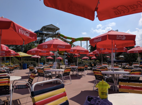 A vibrant water park area with colorful umbrellas, tables, and a water slide in the background under a sunny sky.