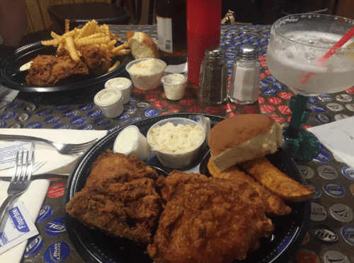 Two plates of fried chicken with sides of coleslaw, fries, and a drink on a patterned table.