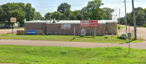 A small, rustic building with signs for Toby's Lounge and Bud Light, surrounded by grass and trees.