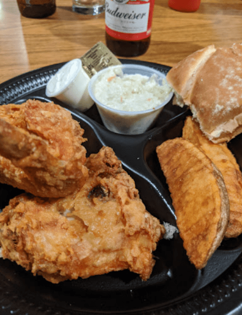 A plate with fried chicken, potato wedges, coleslaw, a roll, and a bottle of Budweiser in the background.