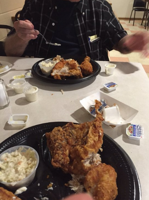 A table set with plates of fried chicken, coleslaw, and dipping sauces, with hands reaching for the food.