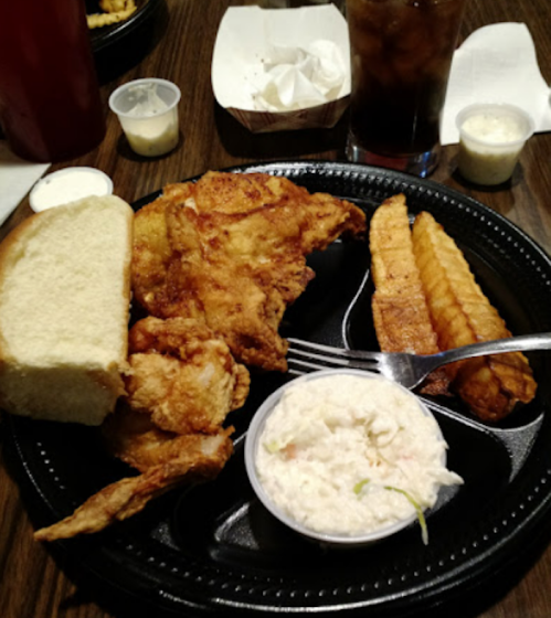 A plate of fried chicken, crinkle-cut fries, coleslaw, a slice of bread, and dipping sauces, with a drink in the background.