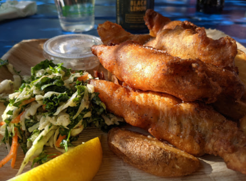 A wooden plate with crispy fried fish, a side of coleslaw, a lemon wedge, and a small cup of dipping sauce.