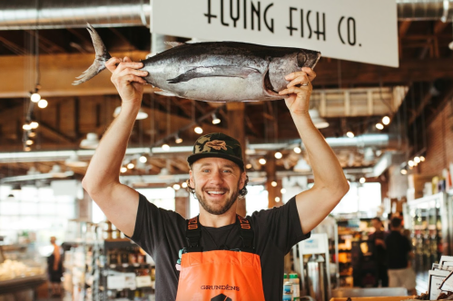 A smiling man in an orange apron holds a large fish above his head in a seafood market.