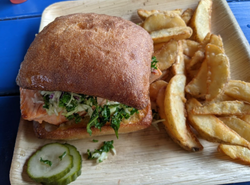 A salmon sandwich with coleslaw on a wooden plate, accompanied by crispy potato wedges and a pickle slice.