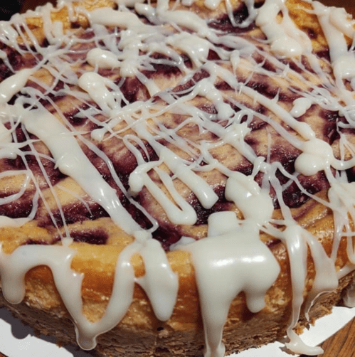 A close-up of a cheesecake with a marbled berry topping and drizzled white icing.