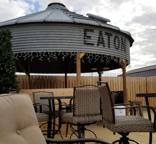 A rustic outdoor seating area with a metal roof structure labeled "EATON," surrounded by wooden fencing and festive lights.