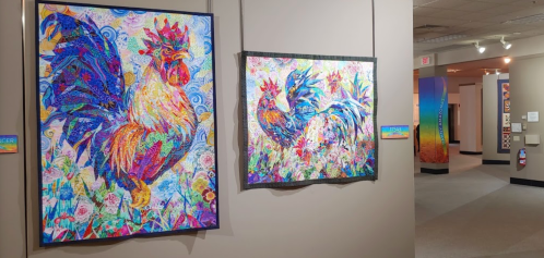 Two vibrant, colorful rooster artworks displayed on a gallery wall, surrounded by a light, neutral background.