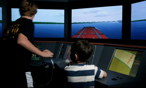 Two children operate a ship simulator, focused on screens displaying water and a bridge in the distance.