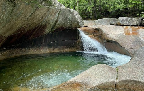 A serene natural scene featuring a small waterfall flowing into a clear, green pool surrounded by smooth rocks and trees.