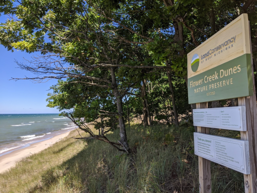 Sign for Flower Creek Dunes Nature Preserve near a sandy beach and trees, with a view of the lake in the background.