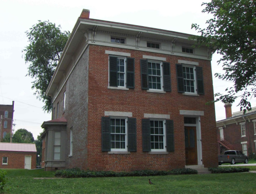 A two-story brick house with black shutters and a green lawn, surrounded by trees and other buildings in the background.