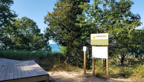 A wooden path leads to a sign for a nature area, with trees and a view of the lake in the background.
