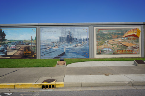 Three colorful murals depicting historical scenes, including cars, industrial sites, and a welcome sign for Atomic City.