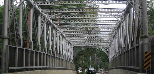 A metal truss bridge with intricate latticework, leading over a road surrounded by trees.