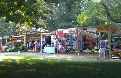 A vibrant outdoor market with wooden stalls selling produce and crafts, surrounded by trees and visitors browsing.