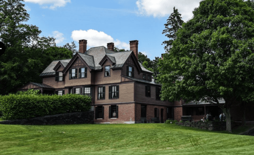 A large, historic house with a brown exterior, surrounded by green grass and trees under a partly cloudy sky.