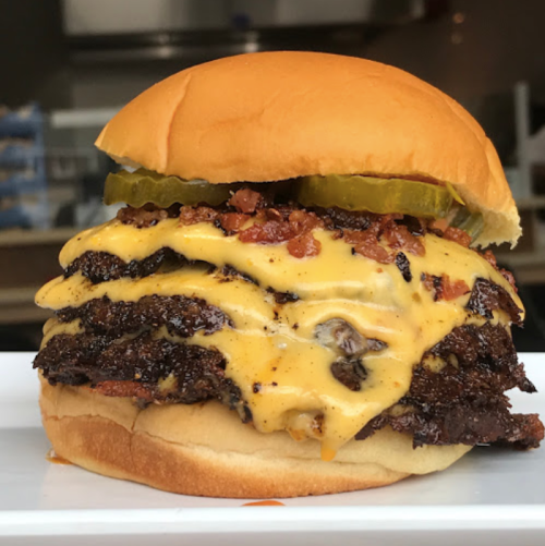 A stacked cheeseburger with melted cheese, bacon, pickles, and a soft bun, served on a white plate.