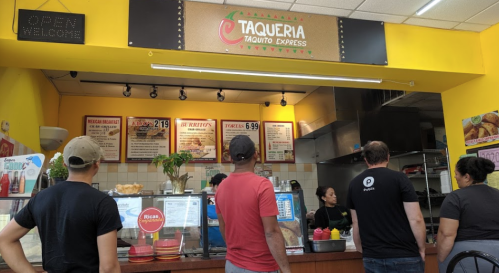 A busy taqueria counter with customers ordering, colorful menu boards, and staff preparing food.