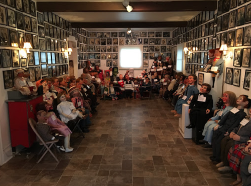 A room filled with vintage dolls and ventriloquist dummies, surrounded by framed photographs on the walls.