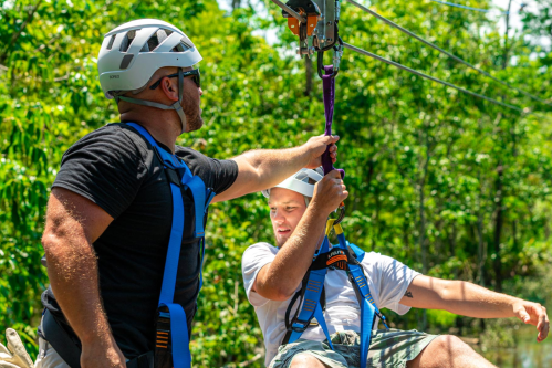 Two men in helmets and harnesses prepare for a zip line ride in a lush, green outdoor setting.
