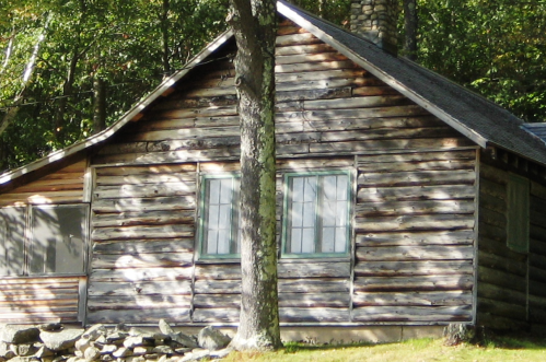 A rustic wooden cabin with a sloped roof, surrounded by trees and dappled sunlight.