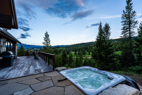 A hot tub on a stone patio overlooks a forested landscape and mountains under a blue sky.