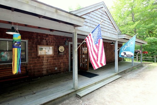 A wooden building with a porch, featuring an American flag and an "Open" banner, surrounded by greenery.
