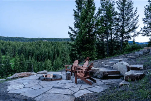 A serene outdoor seating area with wooden chairs and a fire pit, surrounded by lush green trees and a distant forest view.