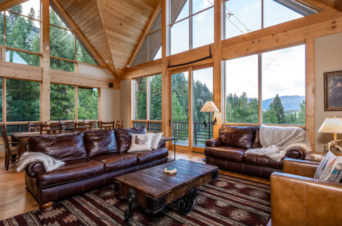 Cozy living room with leather sofas, wooden beams, and large windows showcasing a forest view.