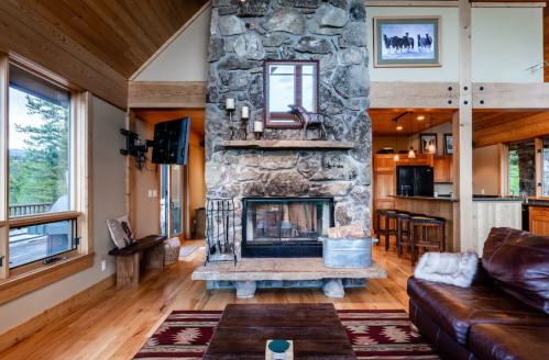 Cozy living room with a stone fireplace, wooden accents, and large windows overlooking nature.