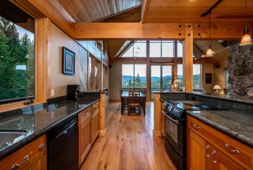 Modern kitchen with wooden cabinets, granite countertops, and large windows showcasing a mountain view. Cozy dining area visible.