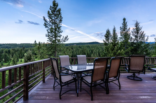 A wooden deck with a glass table and chairs overlooks a lush green forest and mountains under a blue sky.